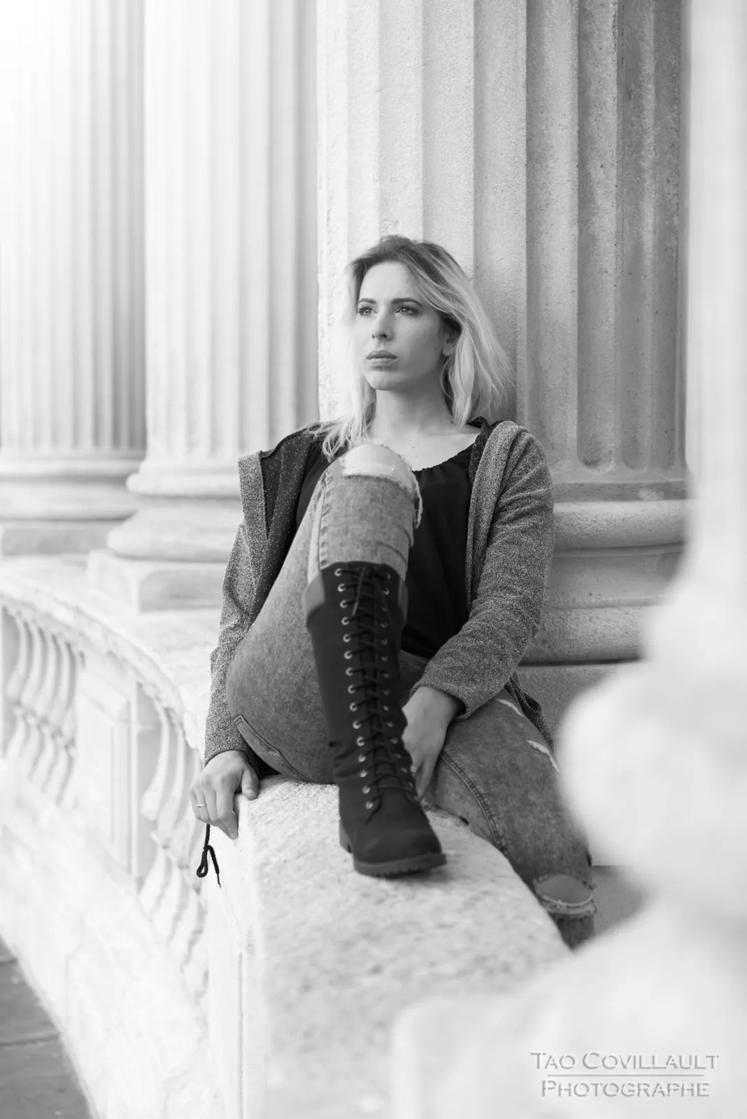 Black-and-white photograph: woman in jeans and a dark jacket posing beside stone columns.