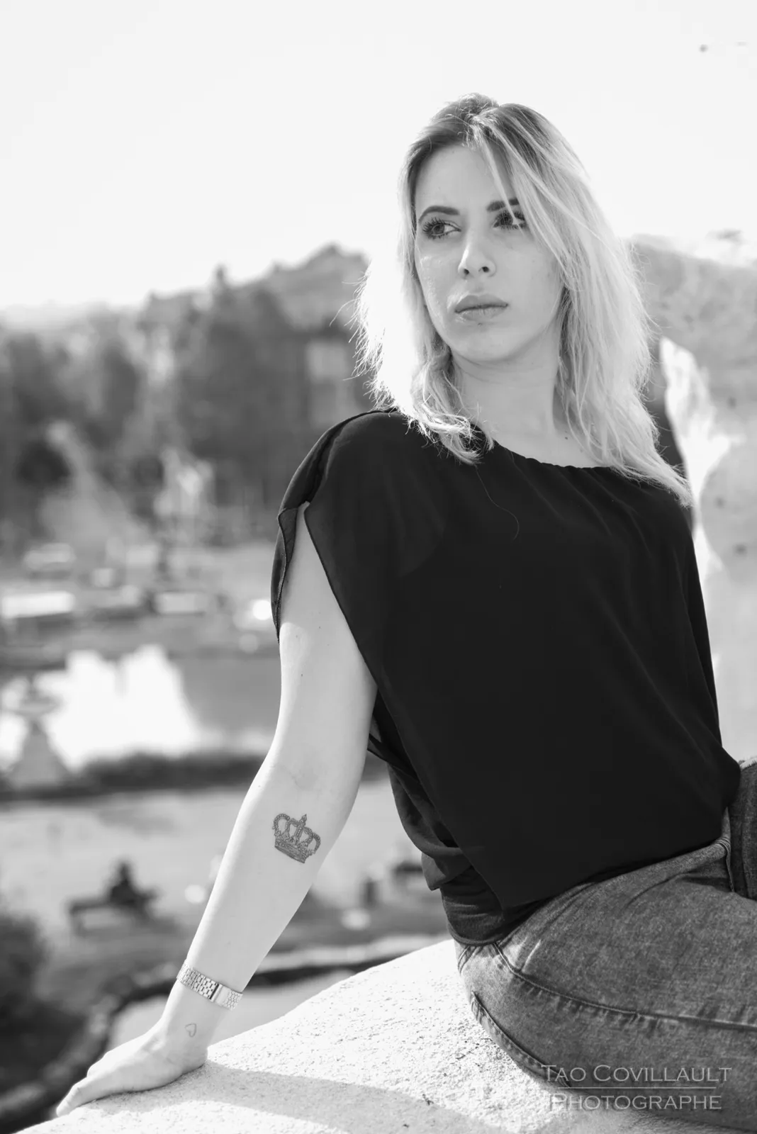 Black-and-white photograph: woman in a black top and jeans seated on a stone balustrade.