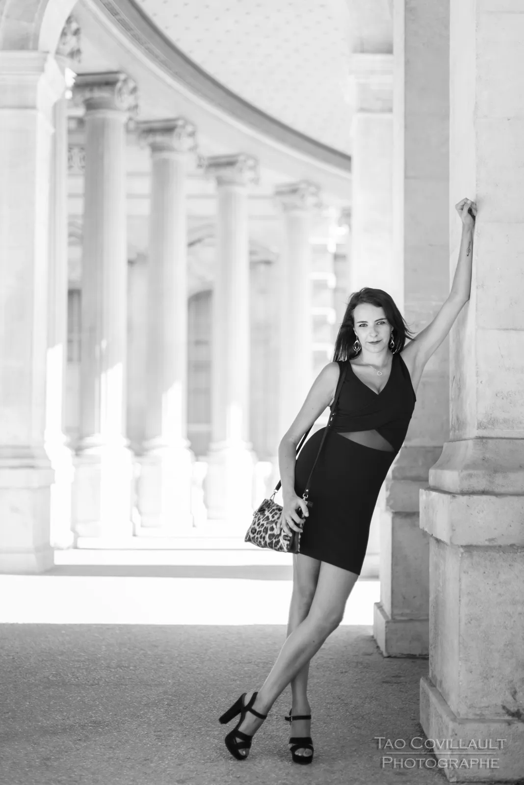 Black-and-white photograph: woman in a black dress posing along a sunlit colonnade.