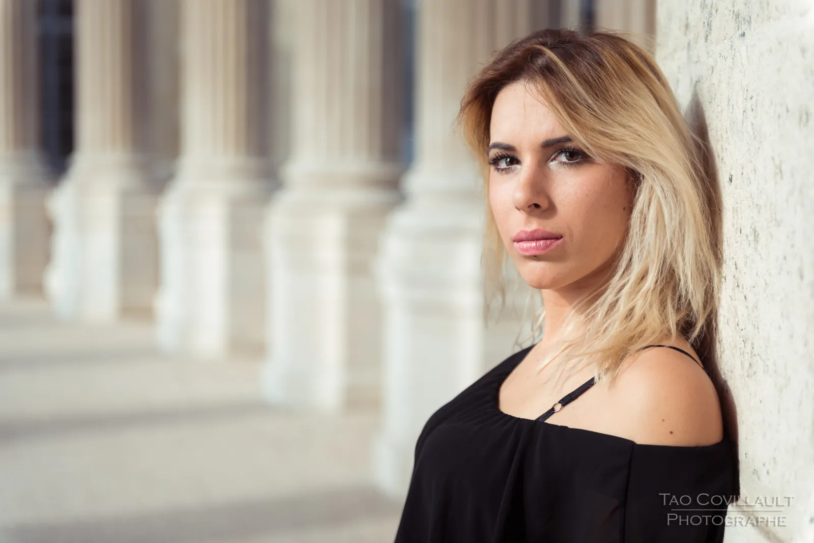Woman in a black top and jeans posing along a stone colonnade.