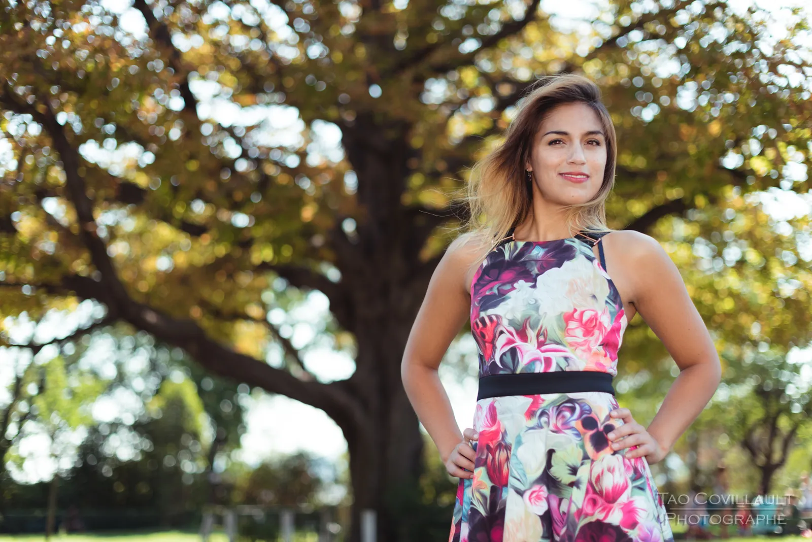 Woman in a floral dress posing beneath leafy trees.