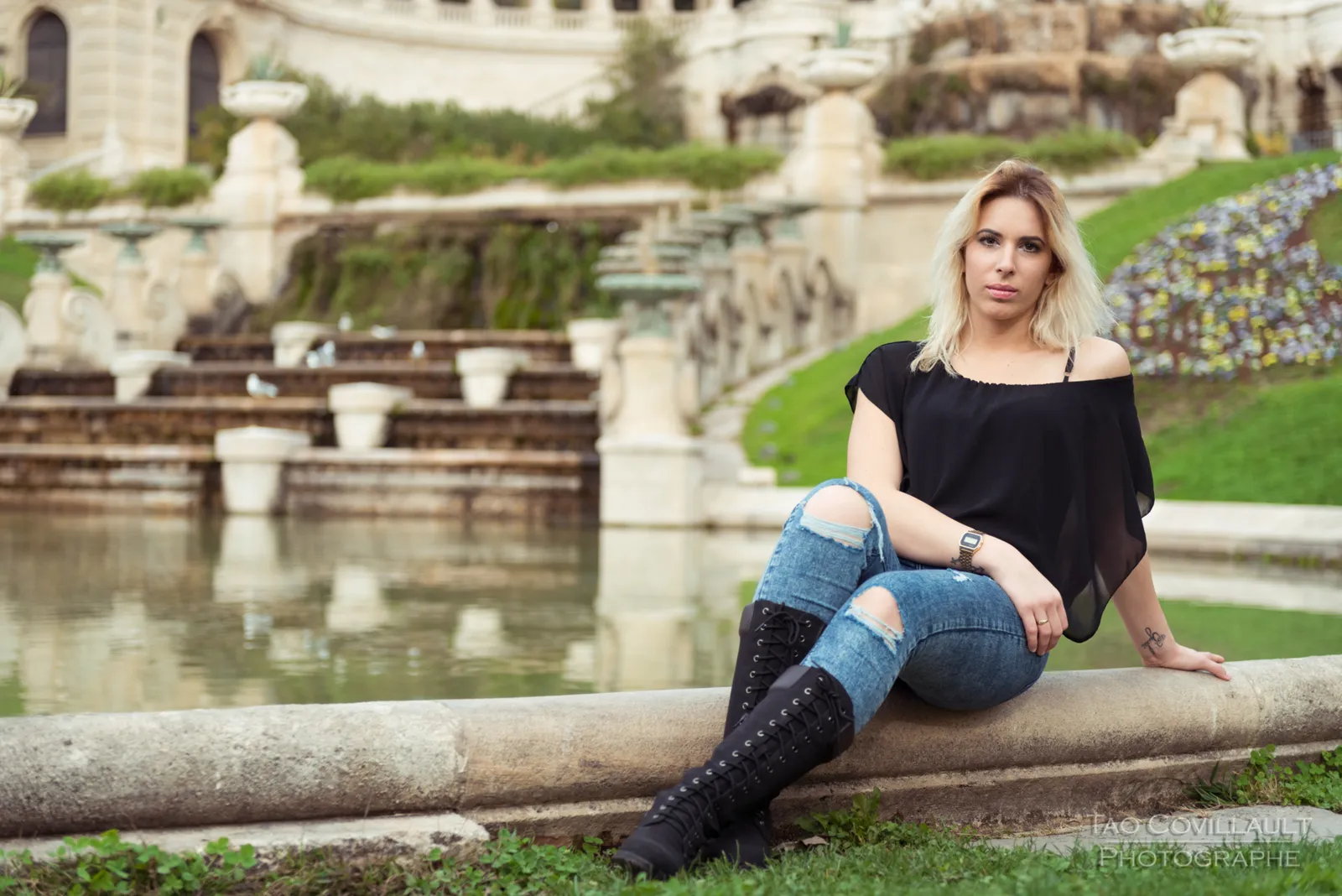 Woman in a black top and jeans sitting on grass beside a flower bed.