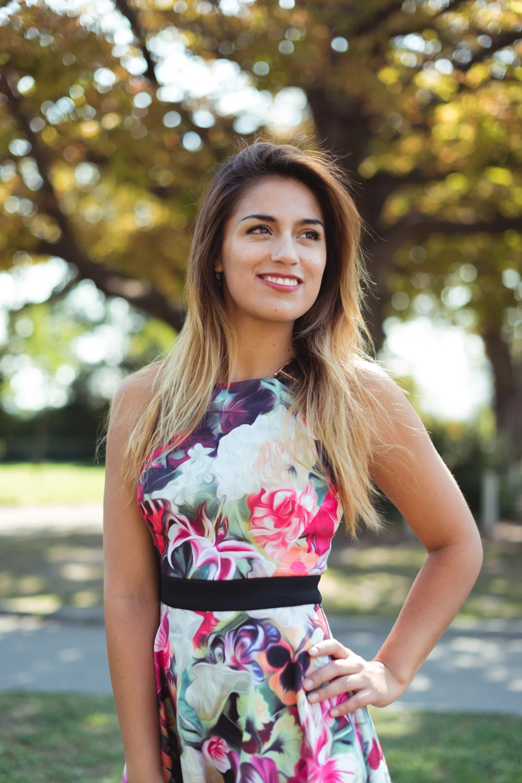Woman in a floral dress posing beneath leafy trees.