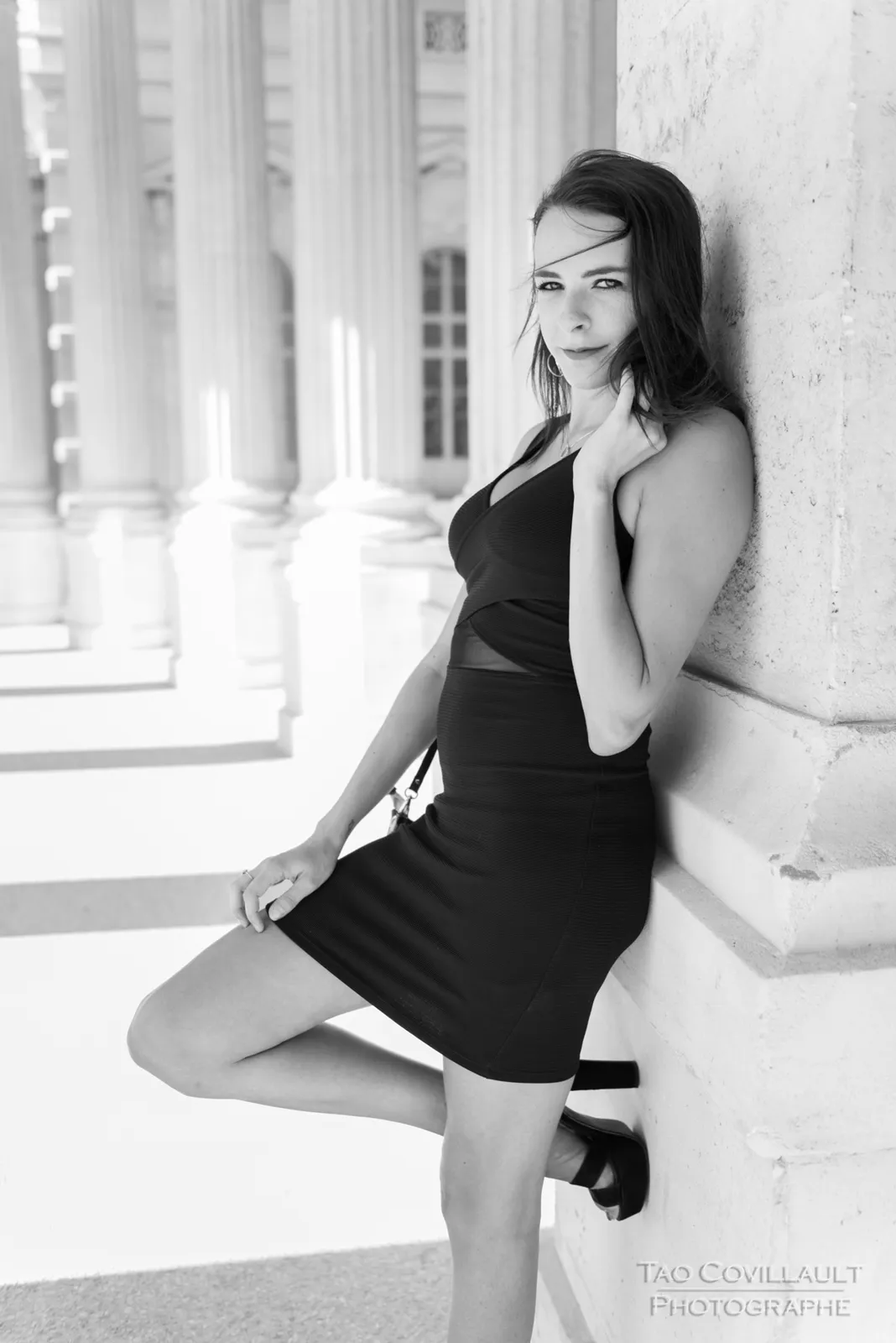 Black-and-white photograph: woman in a black dress posing along a sunlit colonnade.