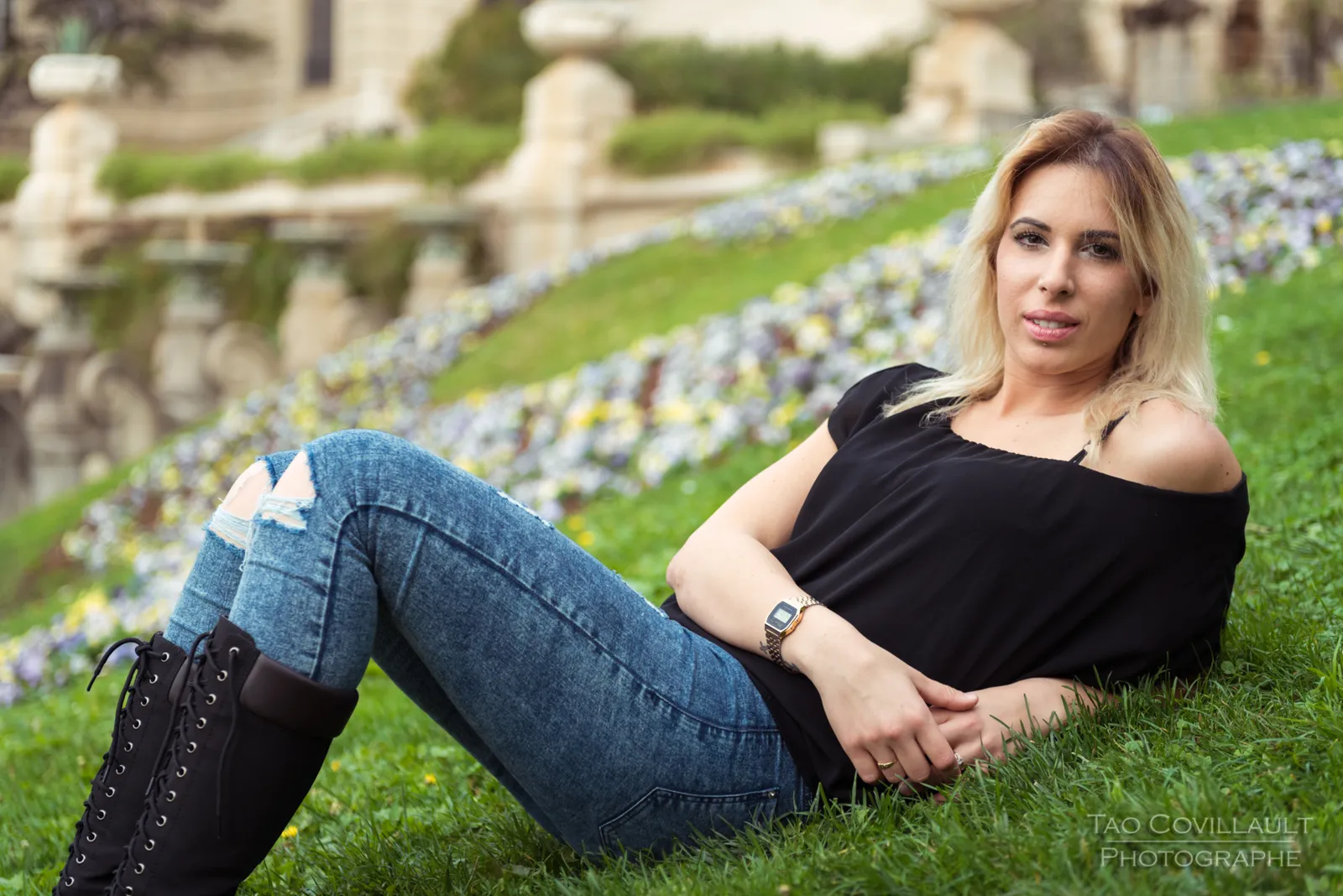 Woman in a black top and jeans sitting on grass beside a flower bed.