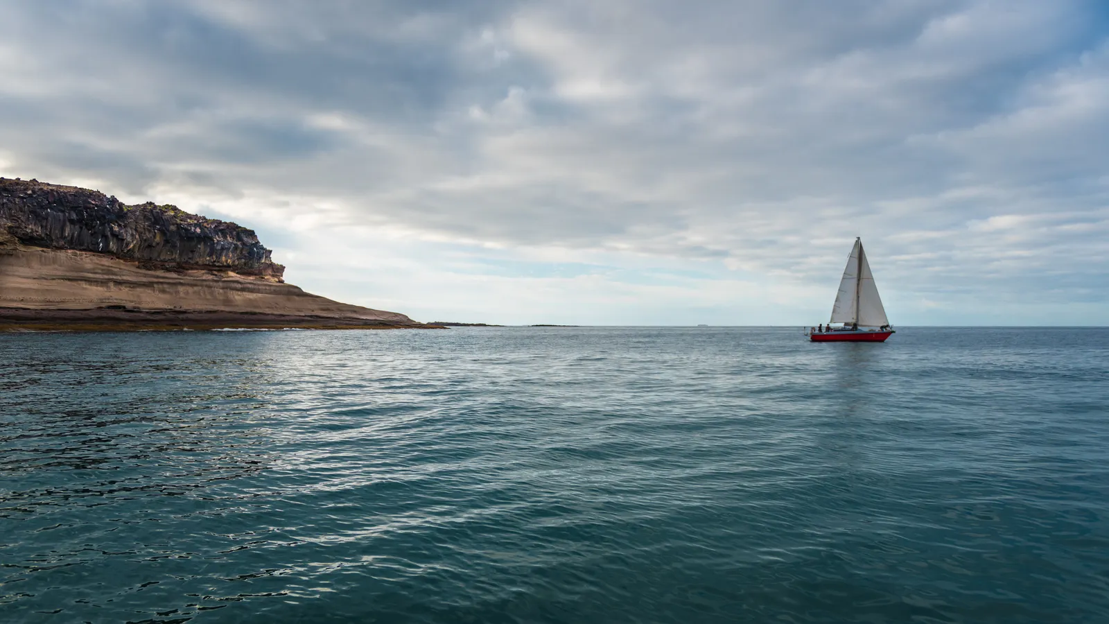 White sailboat off a rocky coastline beneath gray clouds