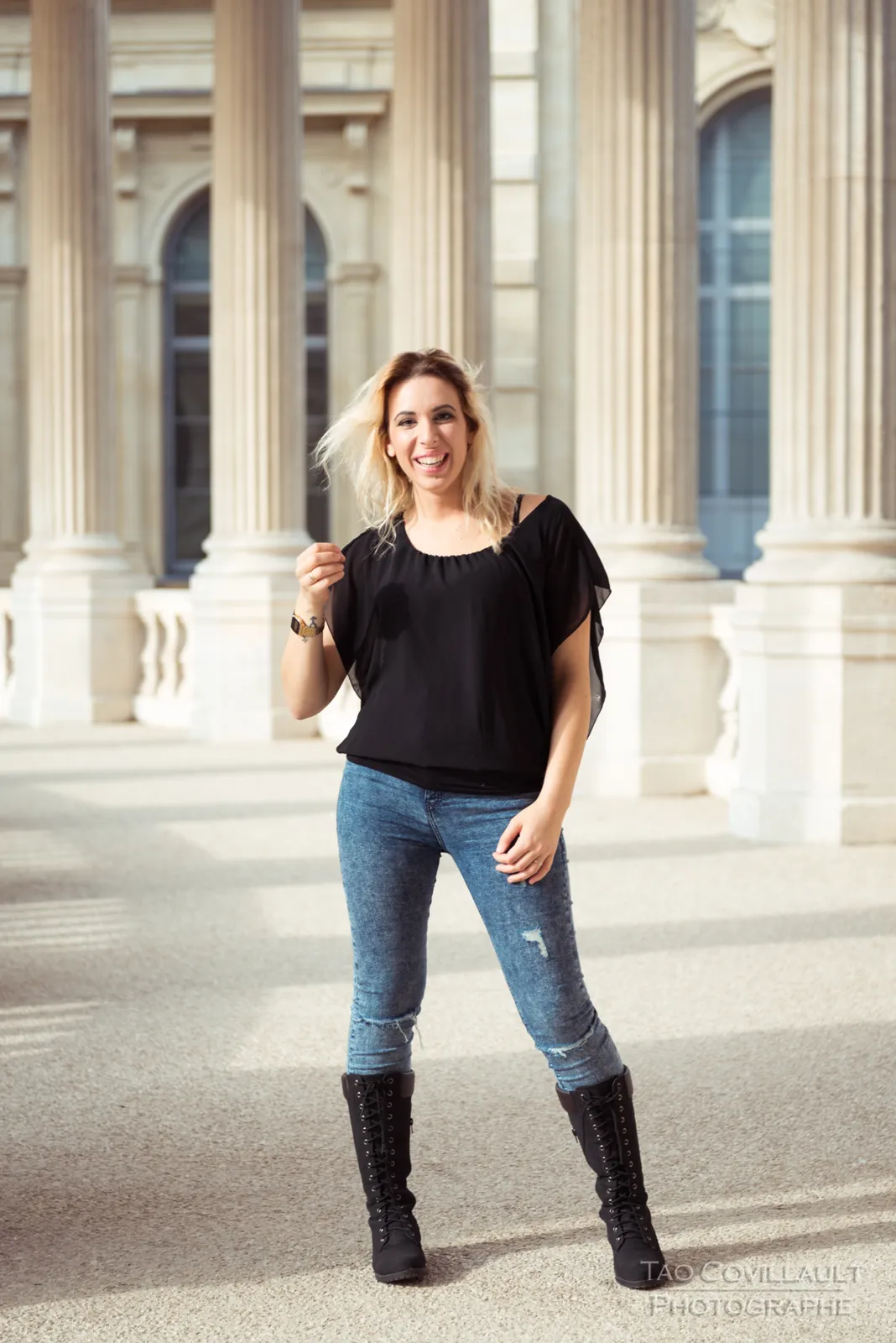 Woman in a black top and jeans posing along a stone colonnade.