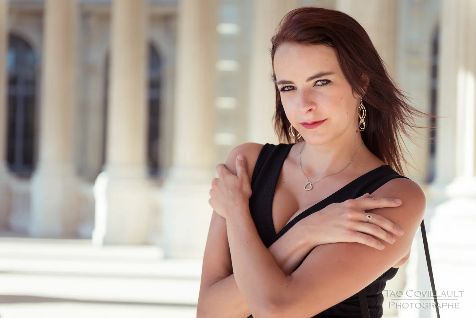 Woman in a black dress posing along a sunlit colonnade.