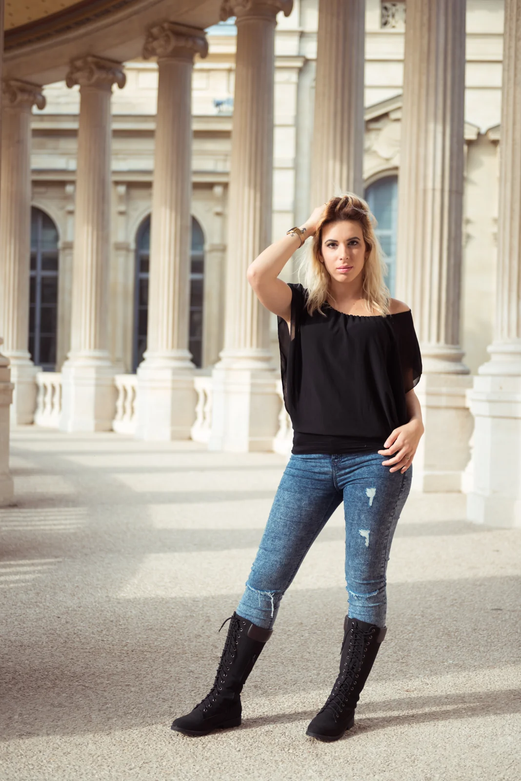 Woman in a black top and jeans posing along a stone colonnade.
