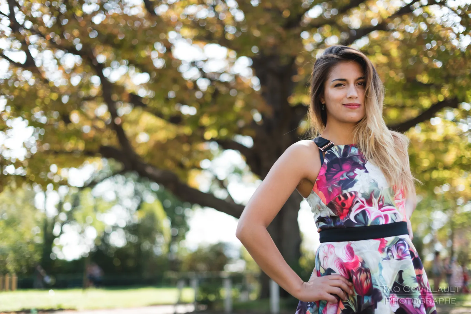Woman in a floral dress posing beneath leafy trees.