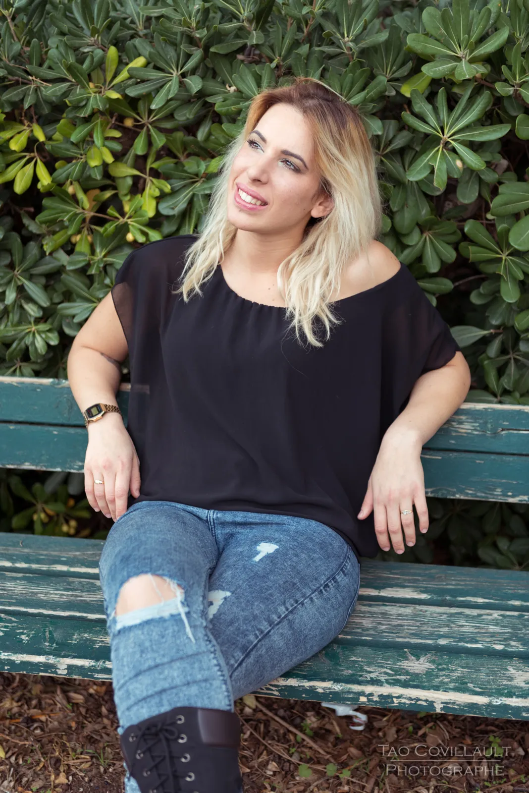Woman in a black top and jeans seated on a green park bench.