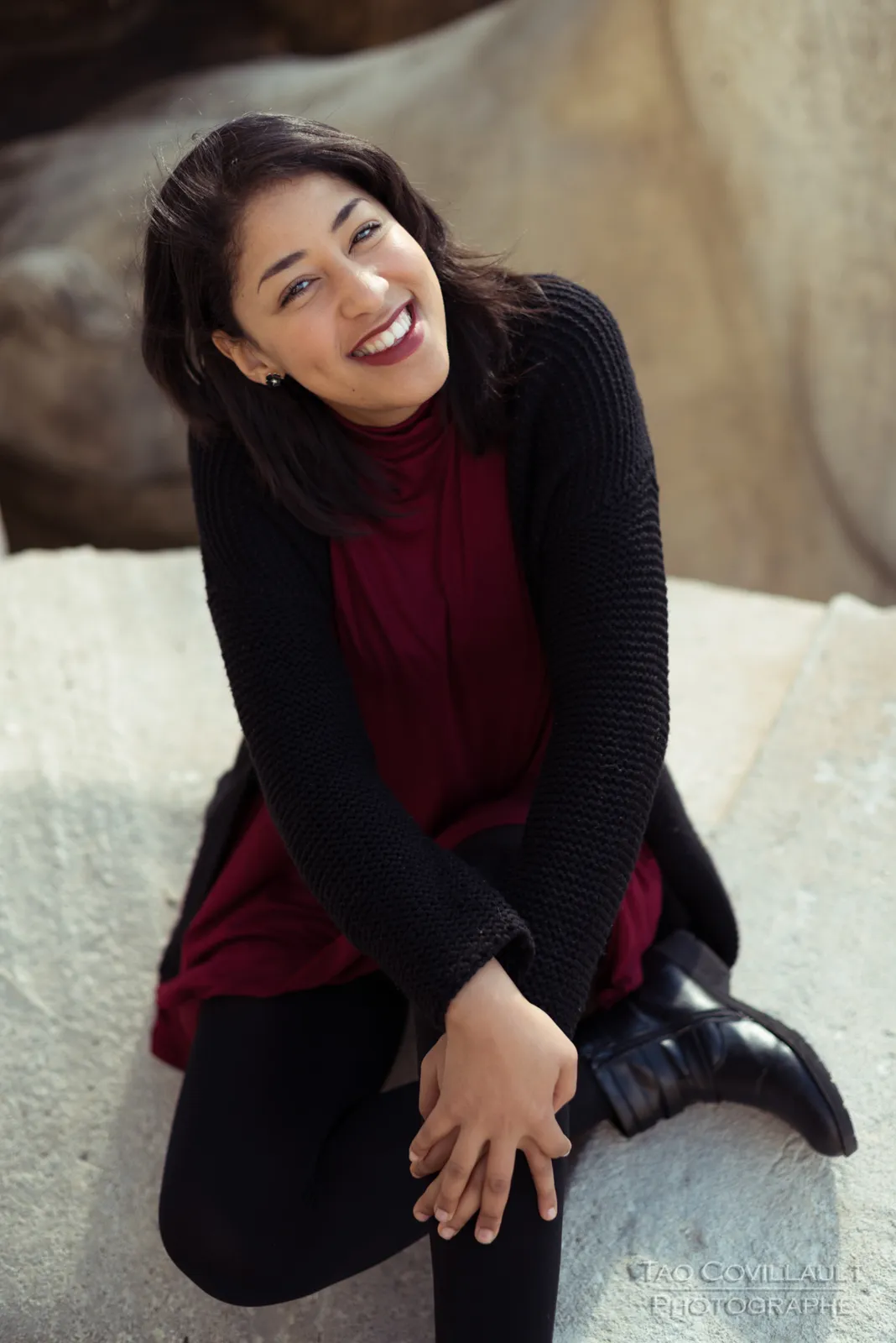 Woman in a burgundy dress and dark coat seated beside a carved stone sculpture.
