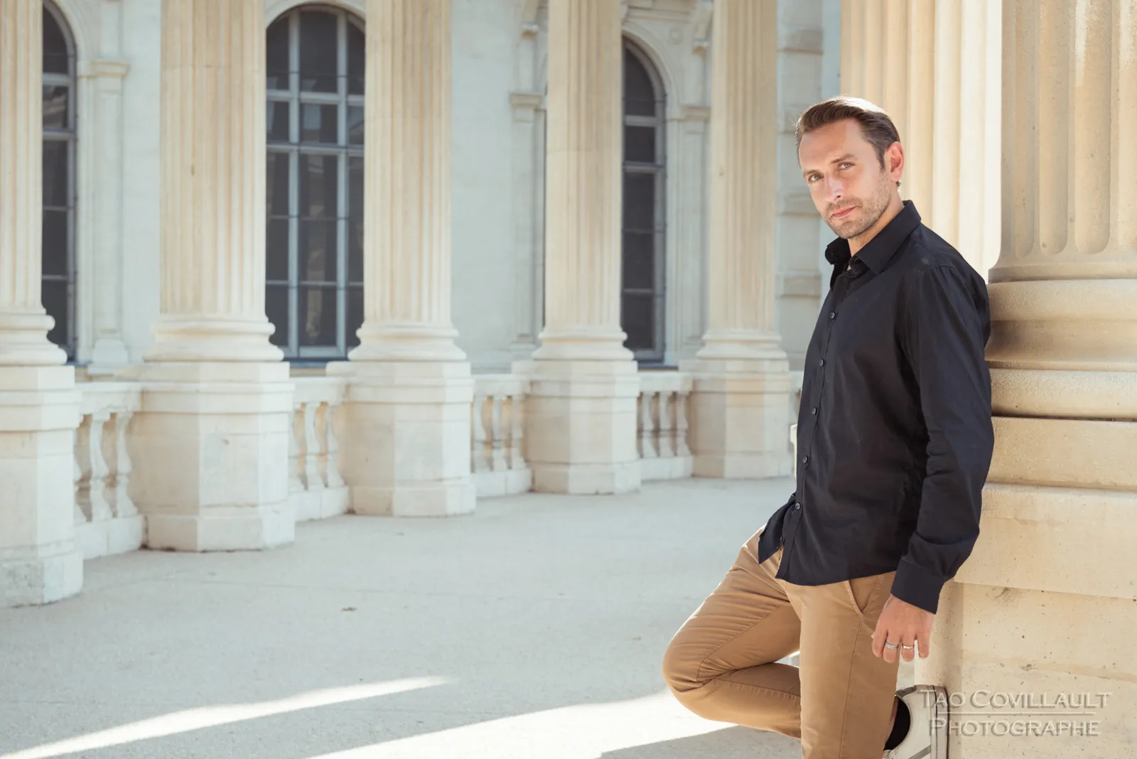 Man in a dark shirt and tan trousers posing in a colonnade.