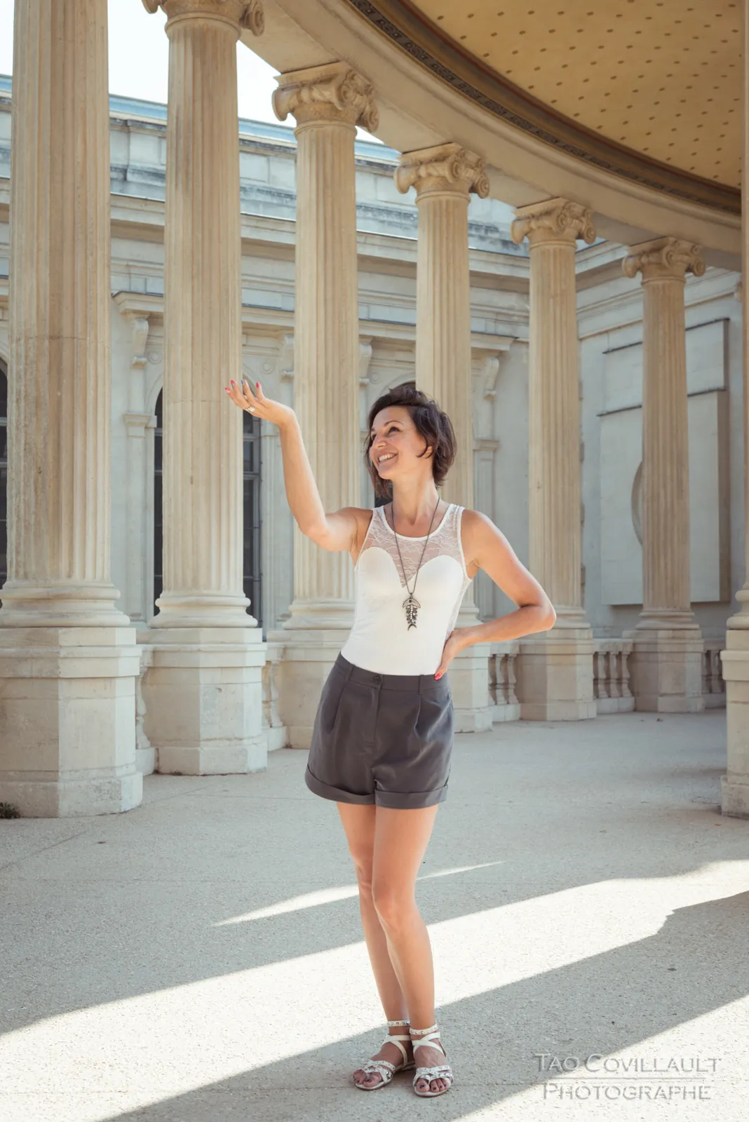 Woman in a white top and dark shorts posing in a colonnade.