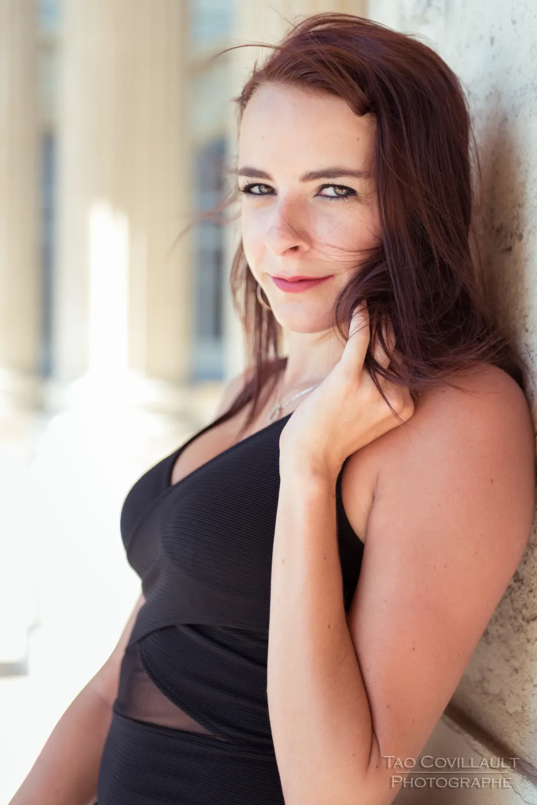 Woman in a black dress posing along a sunlit colonnade.