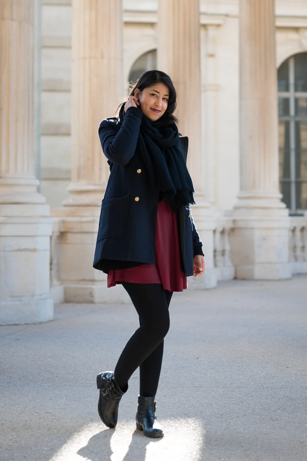 Woman in a dark coat and burgundy dress posing among pale stone columns.