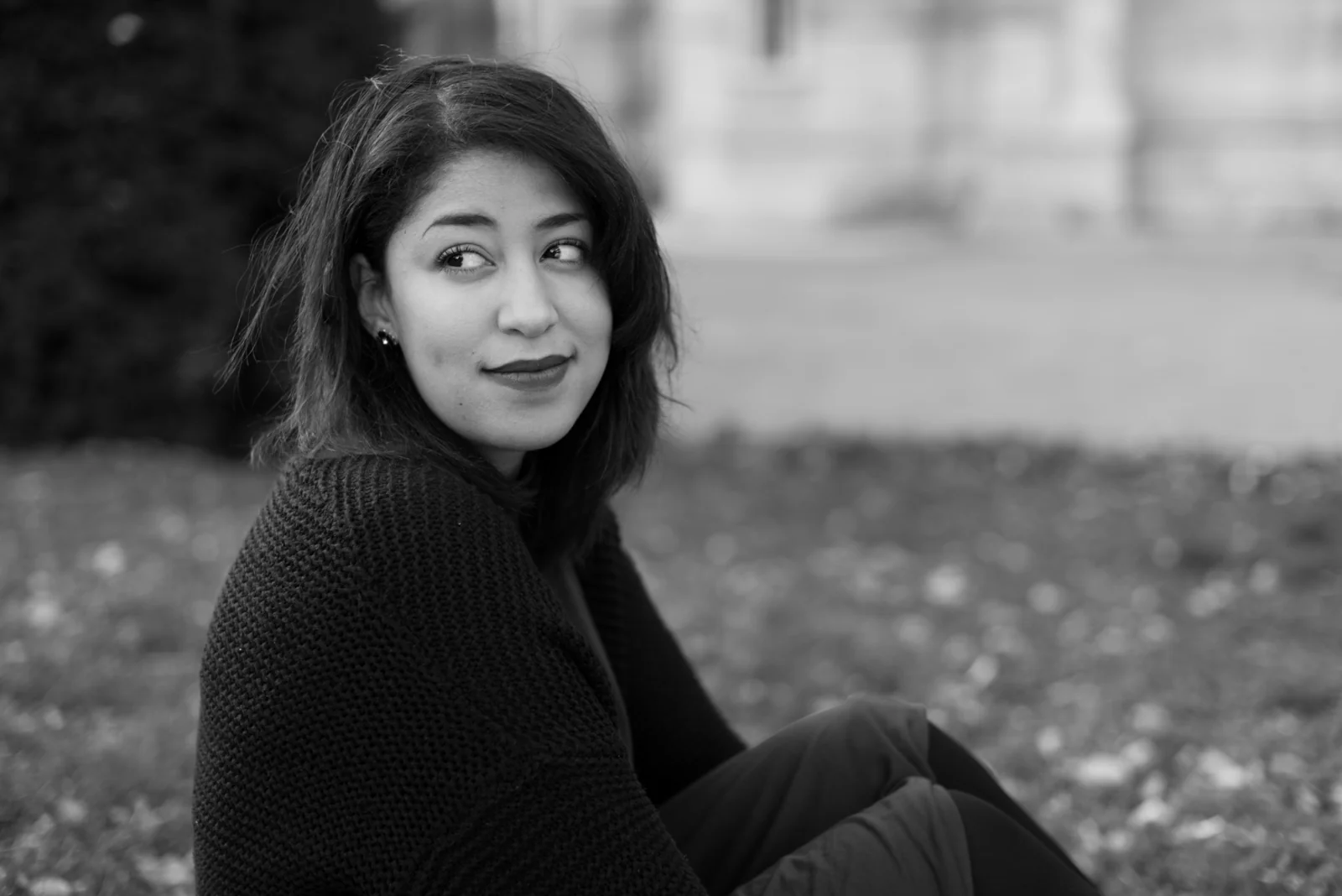Black-and-white photograph: woman in a burgundy dress and dark coat sitting among trees.