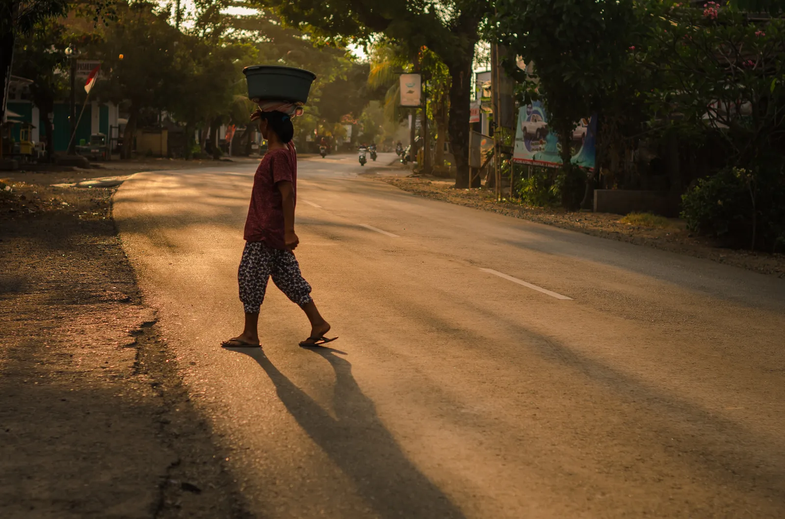 Person crossing a sunlit road with a basket balanced overhead