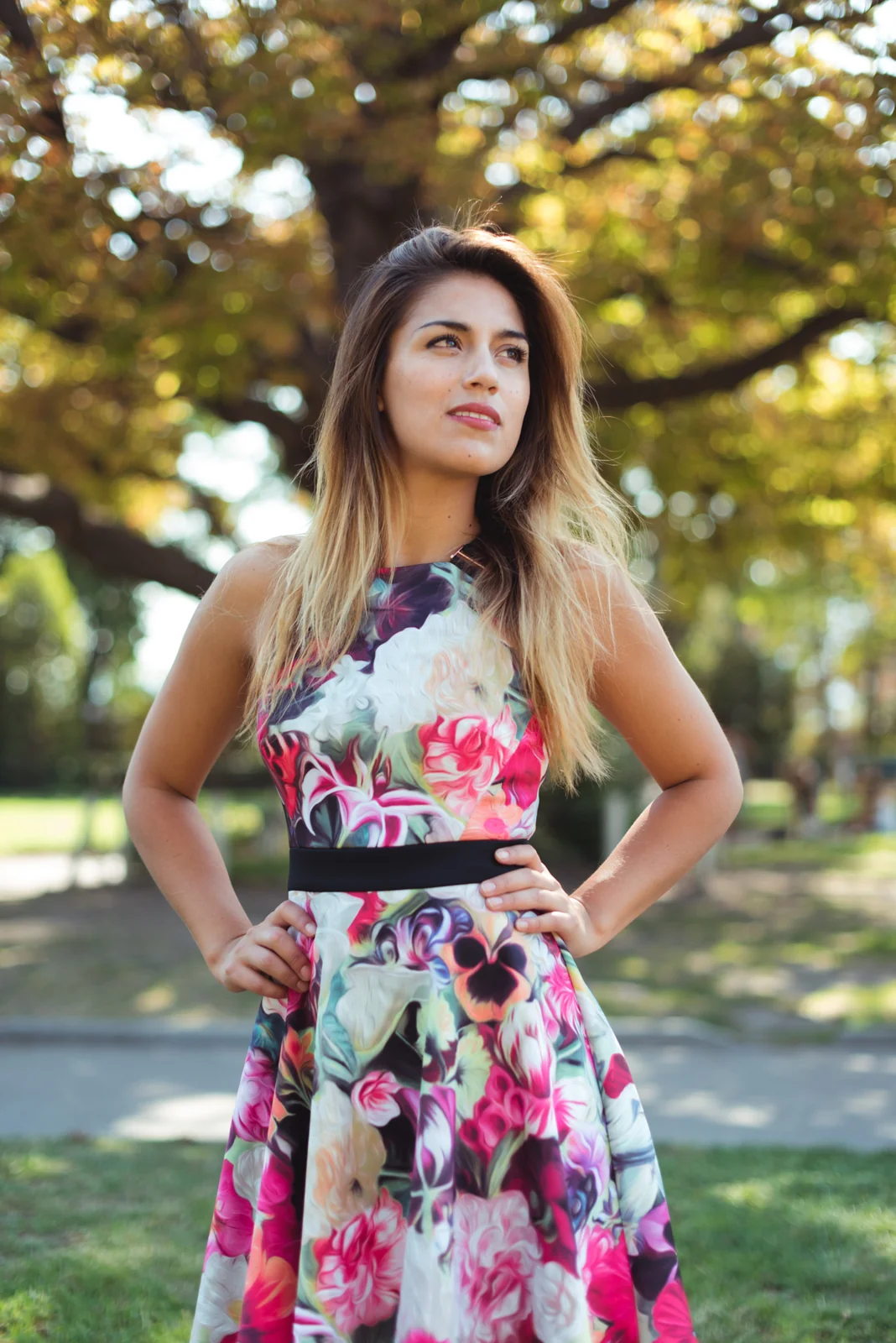 Woman in a floral dress posing beneath leafy trees.