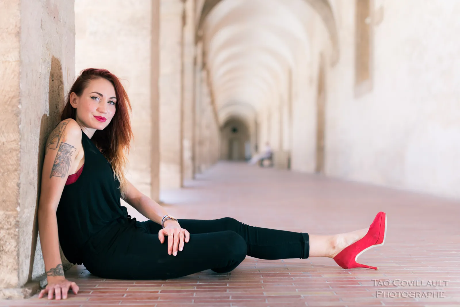 Woman in black clothing and red shoes seated on the floor of a stone arcade.