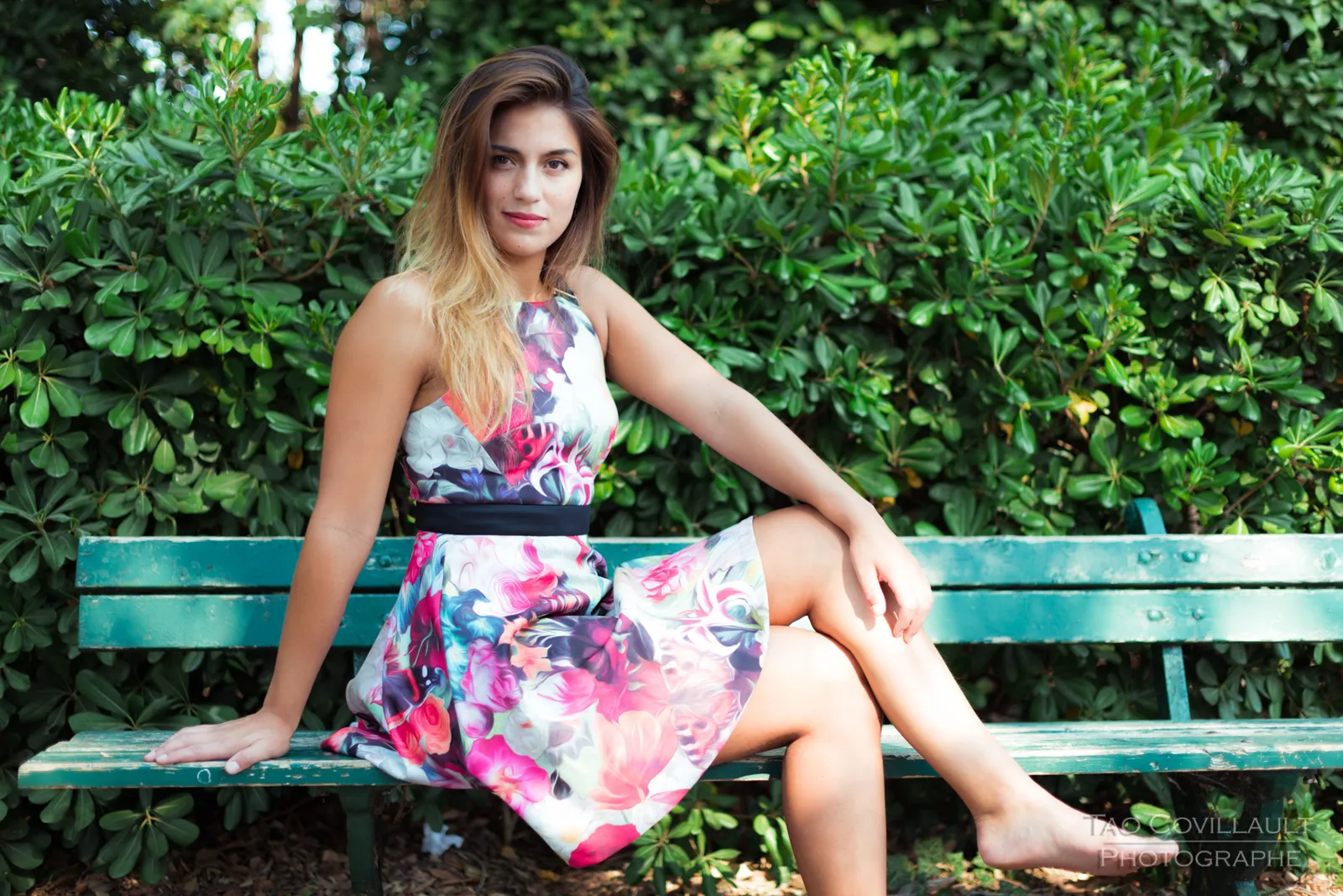 Woman in a floral dress posing beside a green park bench.