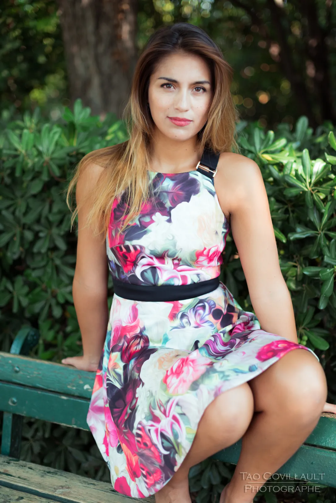 Woman in a floral dress posing beside a green park bench.