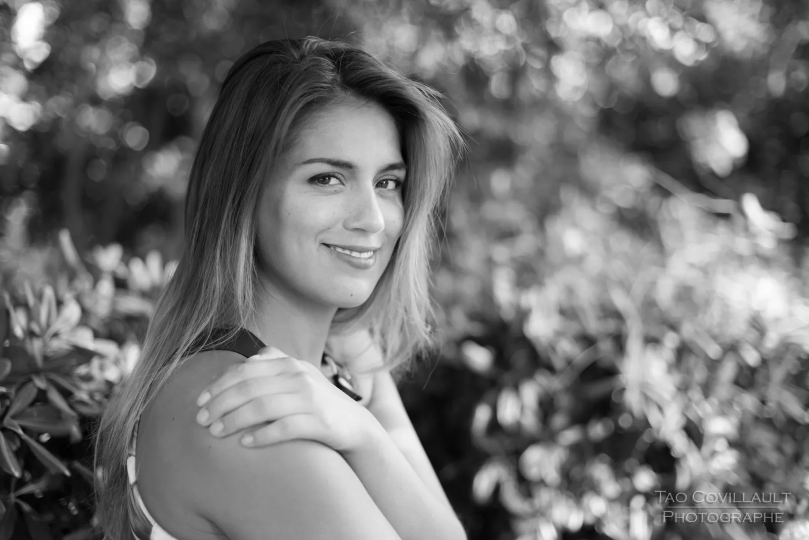 Black-and-white photograph: woman in a floral dress posing beside a green park bench.