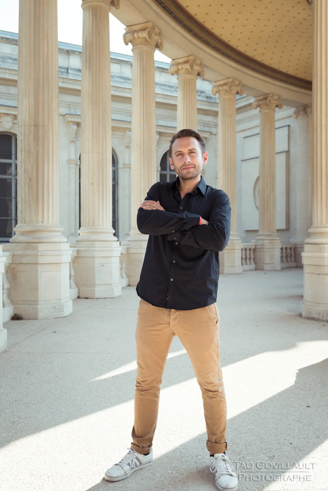 Man in a dark shirt and tan trousers posing in a colonnade.