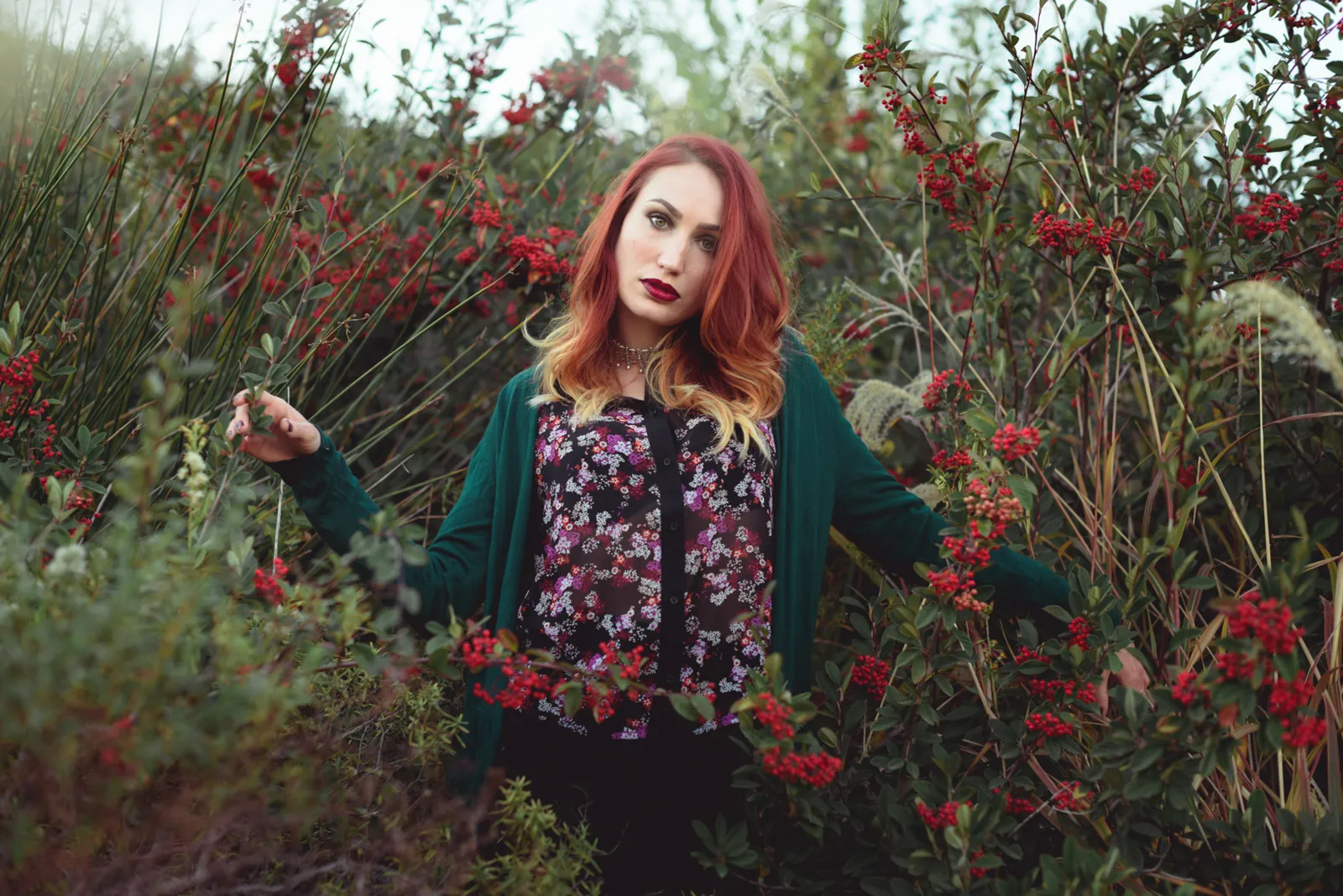 Red-haired woman standing among red flowers and tall branches.