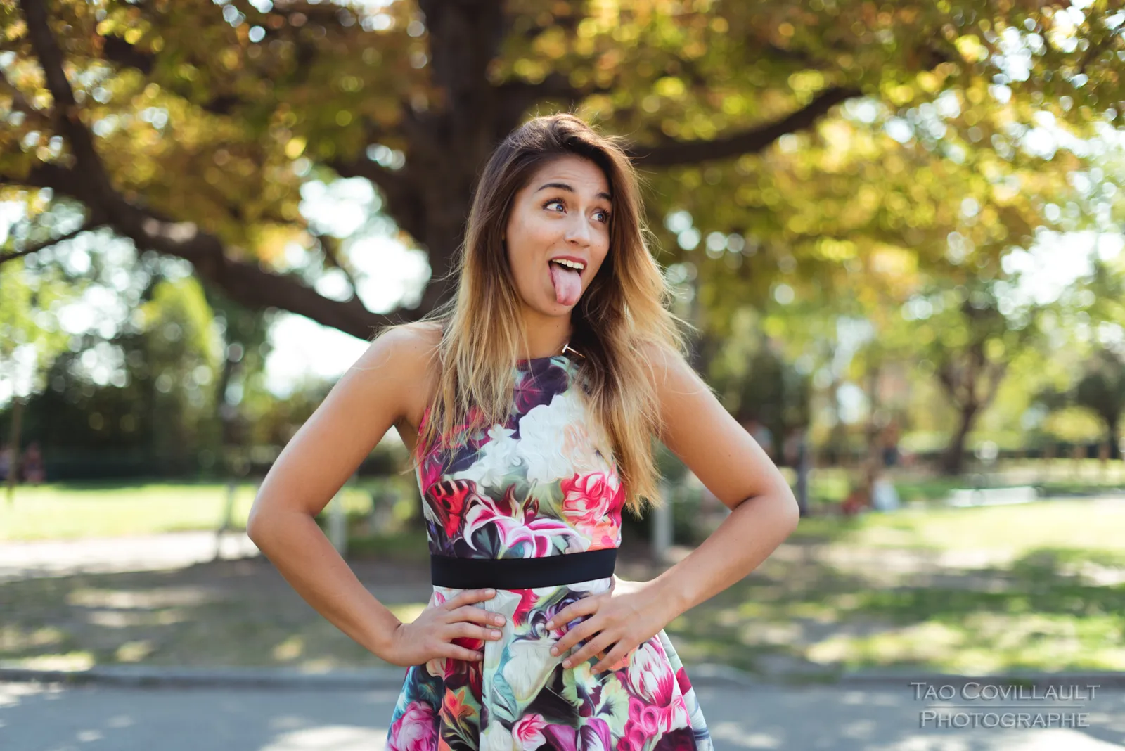 Woman in a floral dress posing beneath leafy trees.
