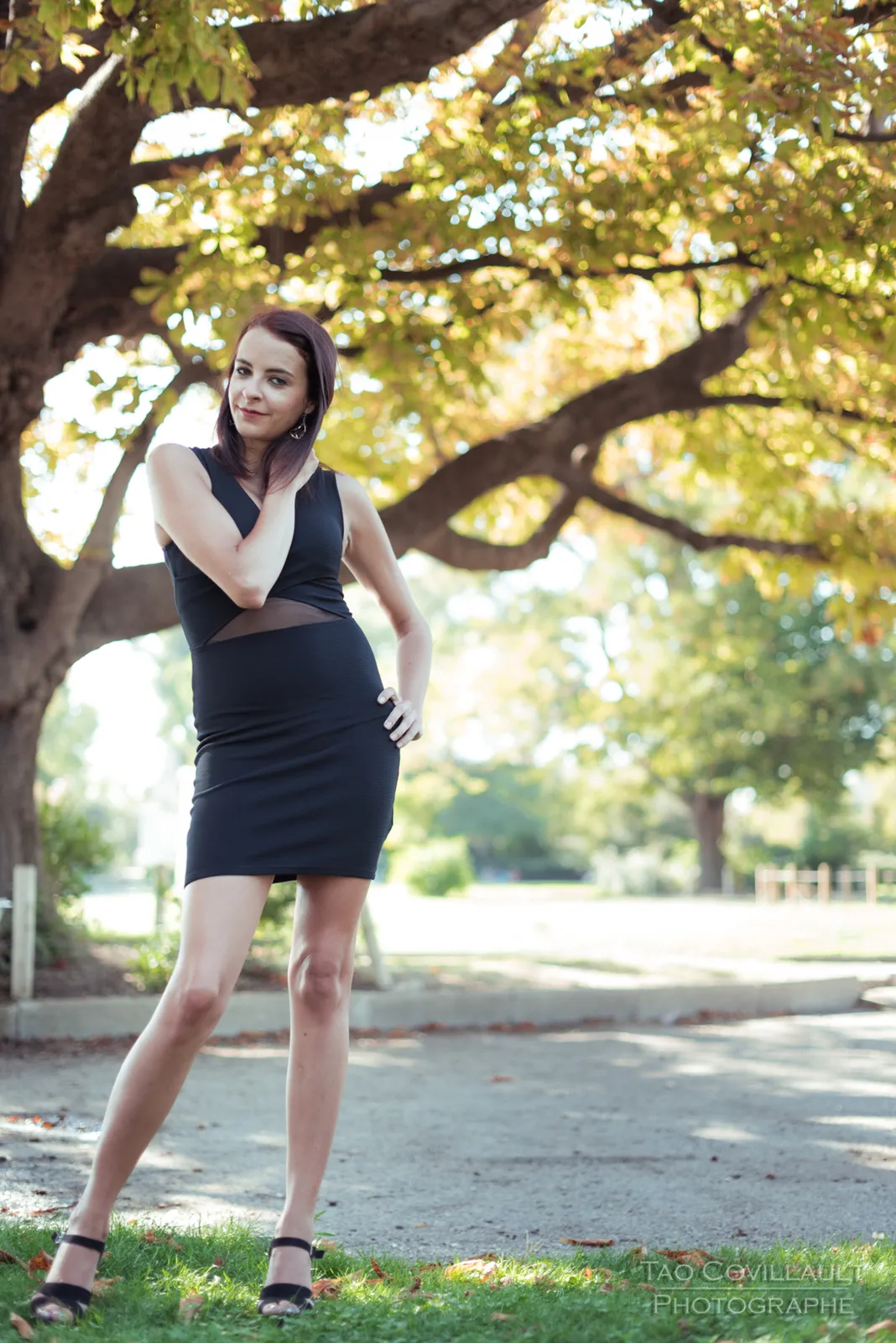 Woman in a black dress standing beneath a leafy tree.