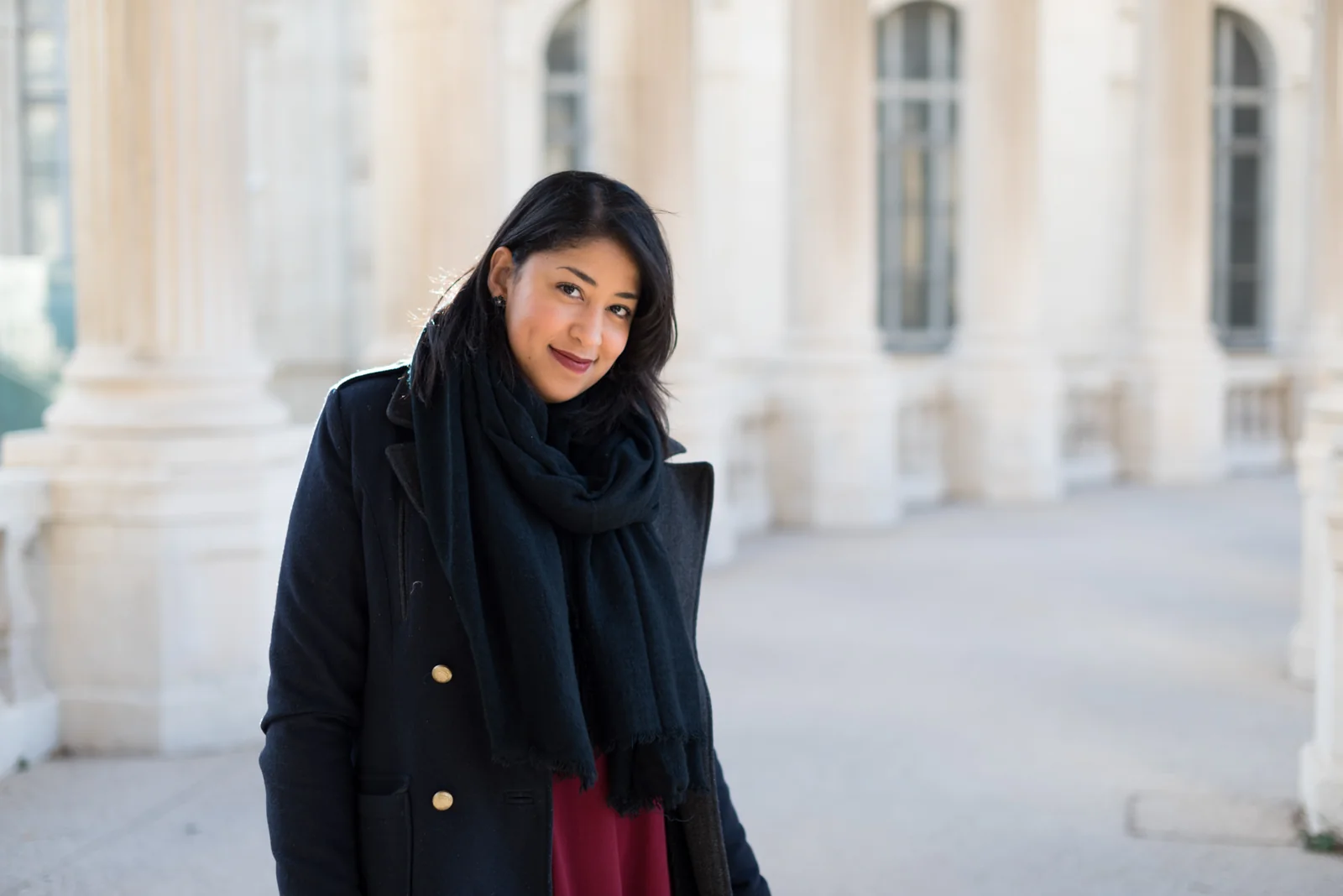 Woman in a dark coat and burgundy dress posing among pale stone columns.