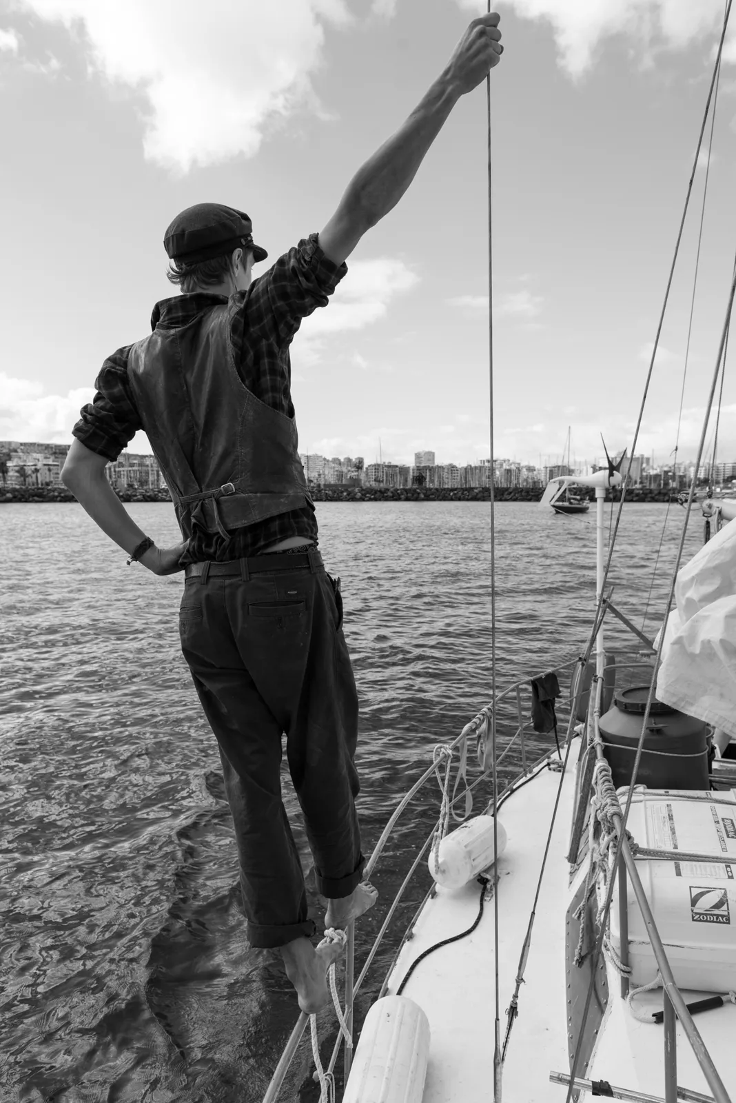 Black-and-white photograph: person standing on a sailboat with one arm raised.