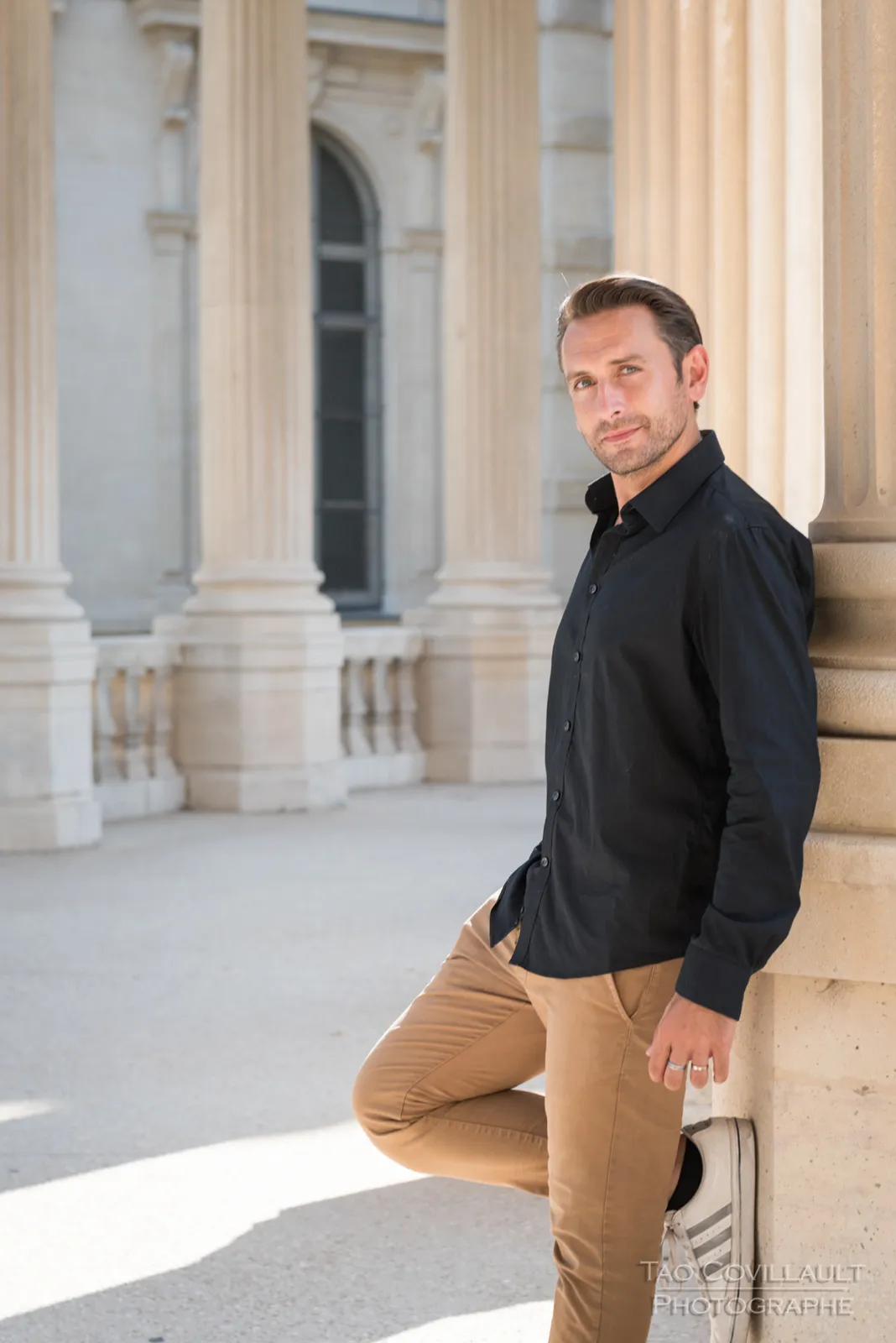 Man in a dark shirt and tan trousers posing in a colonnade.