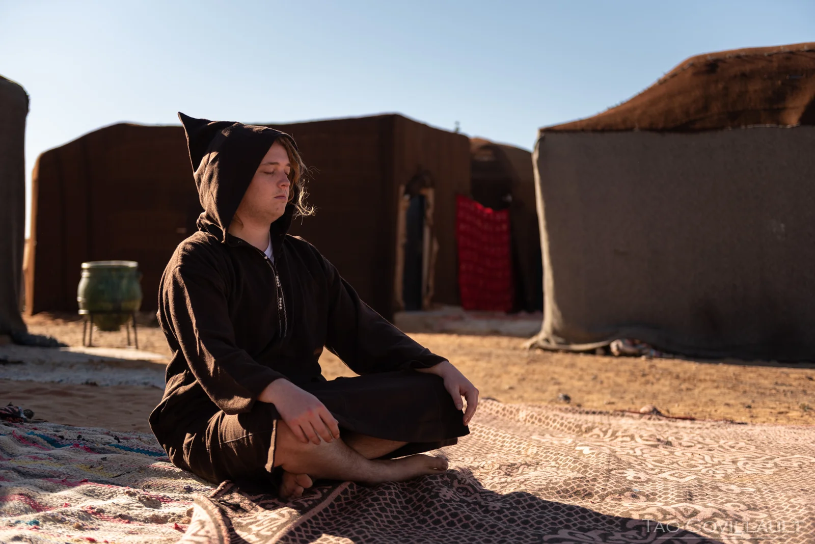 A person sitting cross-legged on a rug outside desert buildings.