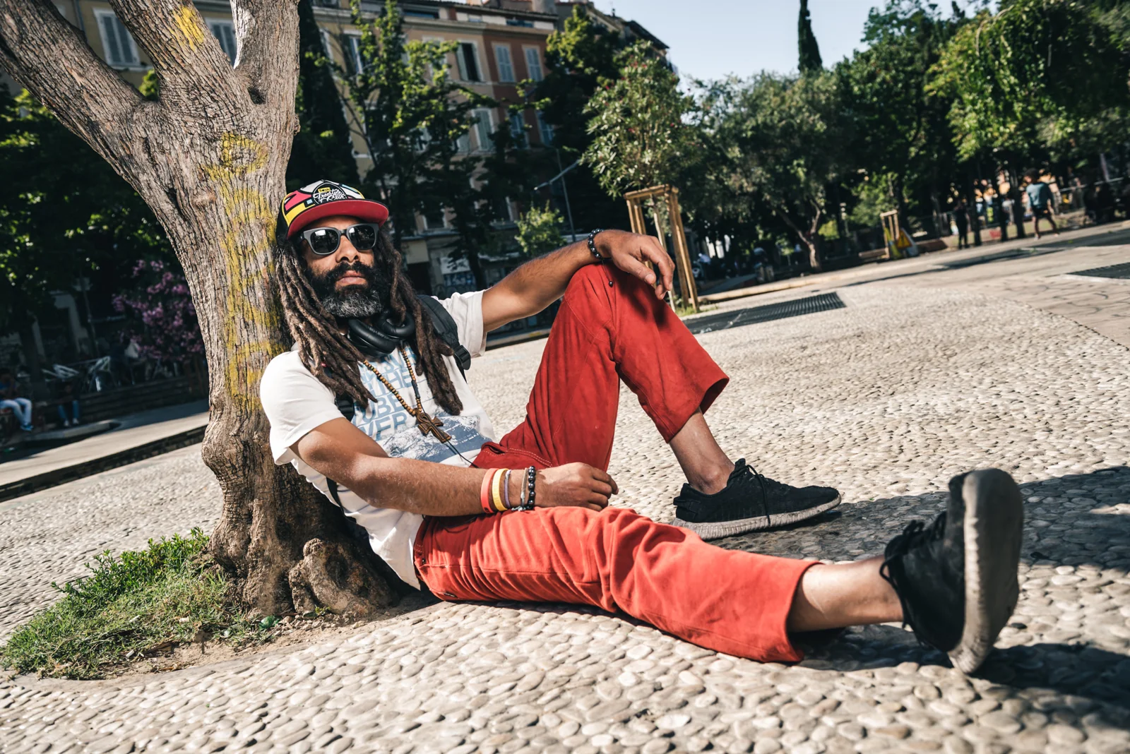 Man with dreadlocks and red trousers reclining beside a tree.