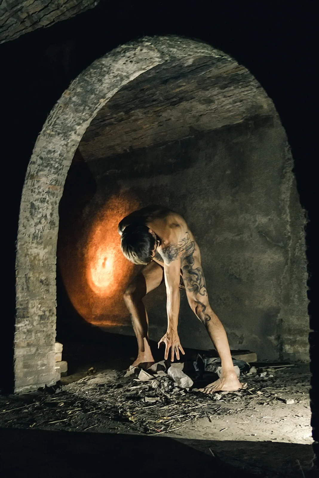 Fine-art nude figure bending toward rubble beneath a stone arch.