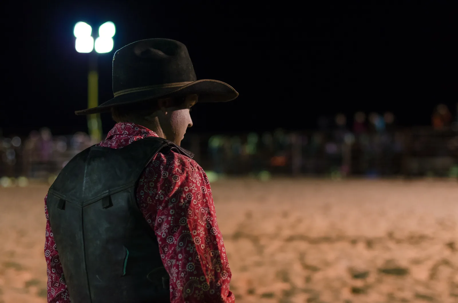 Rodeo participant in a black hat watching the arena at night.