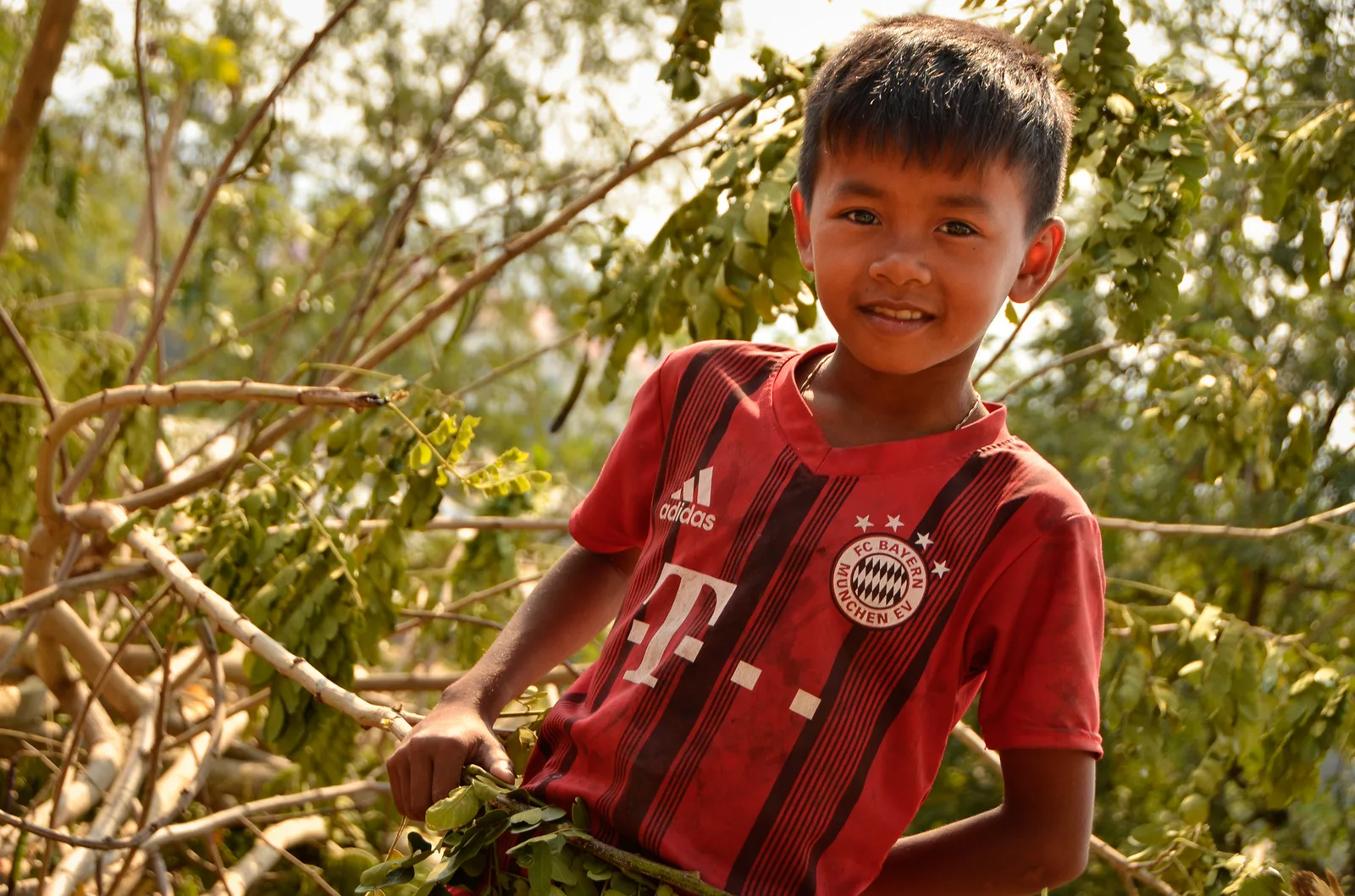 Smiling child in a red football shirt beside leafy branches.