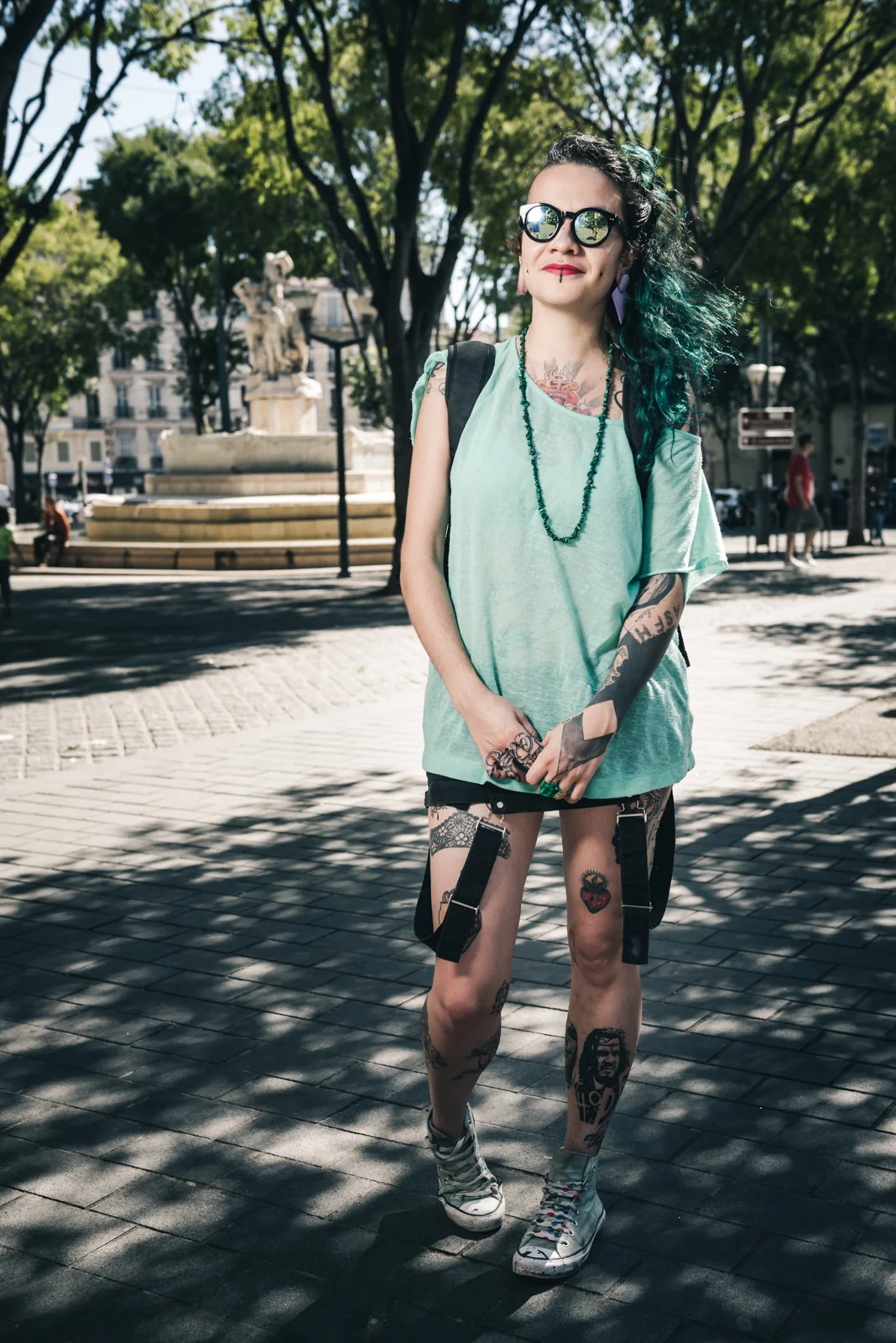 Person with green hair and sunglasses standing in dappled shade.