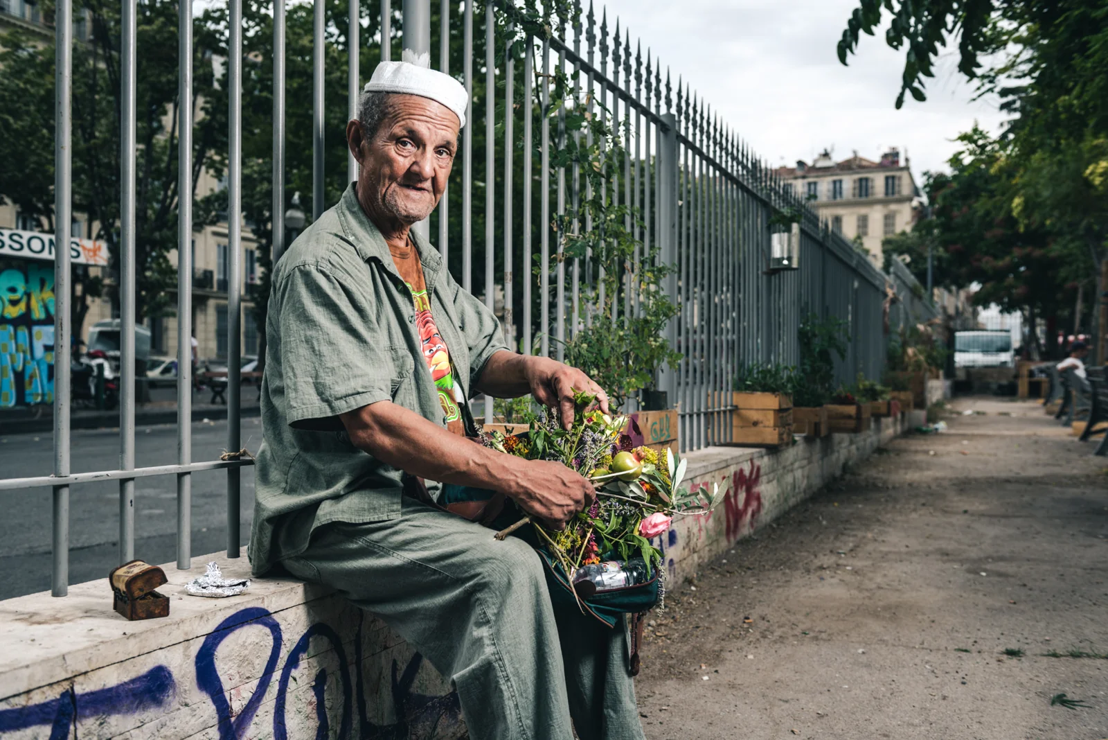Man in pale green clothes seated beside a fence with flowers.