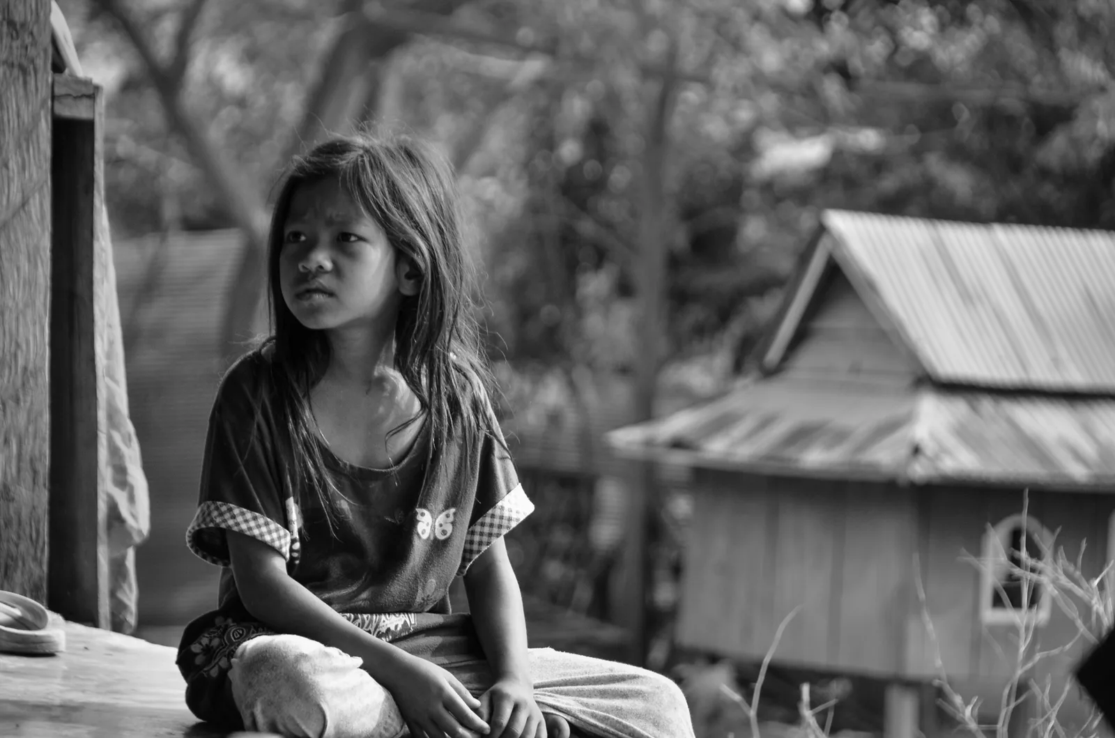 Black-and-white portrait of a child seated near a wooden building.
