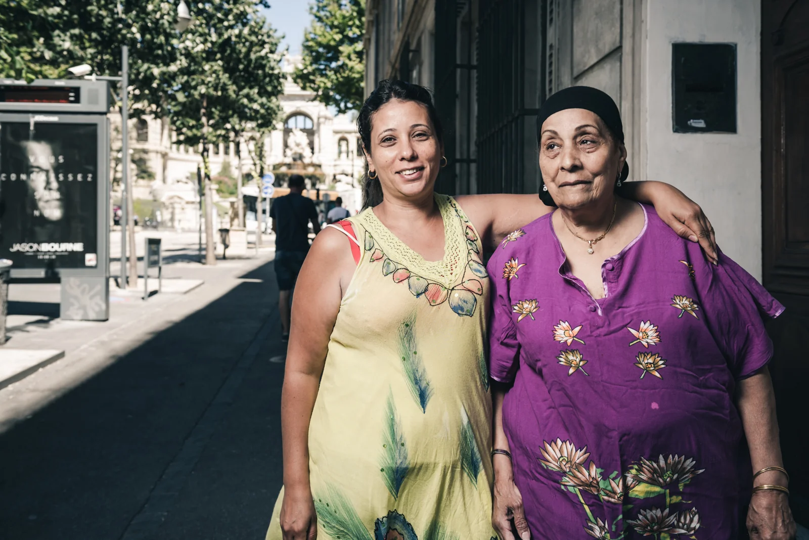 Two women standing arm in arm on a sunlit sidewalk.