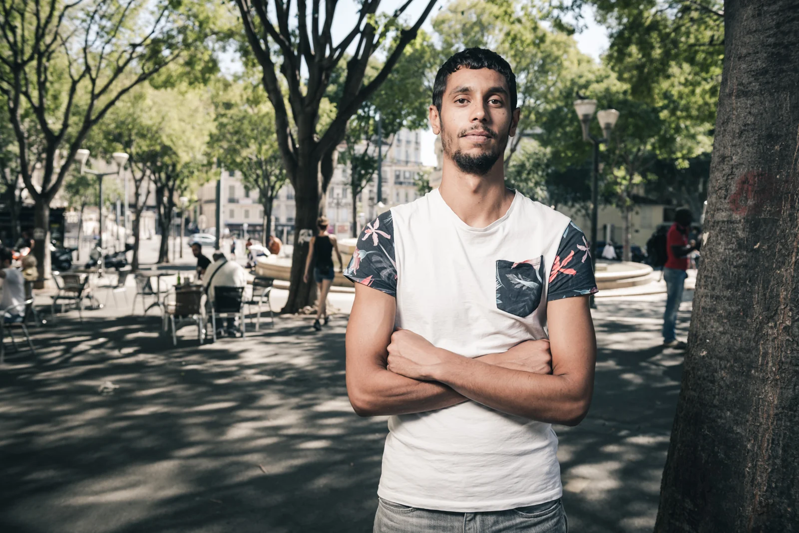 Man in a white T-shirt standing with folded arms in a public square.