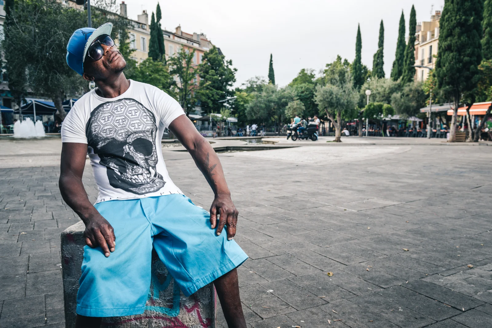 Man in a white T-shirt and blue shorts seated in a city square.