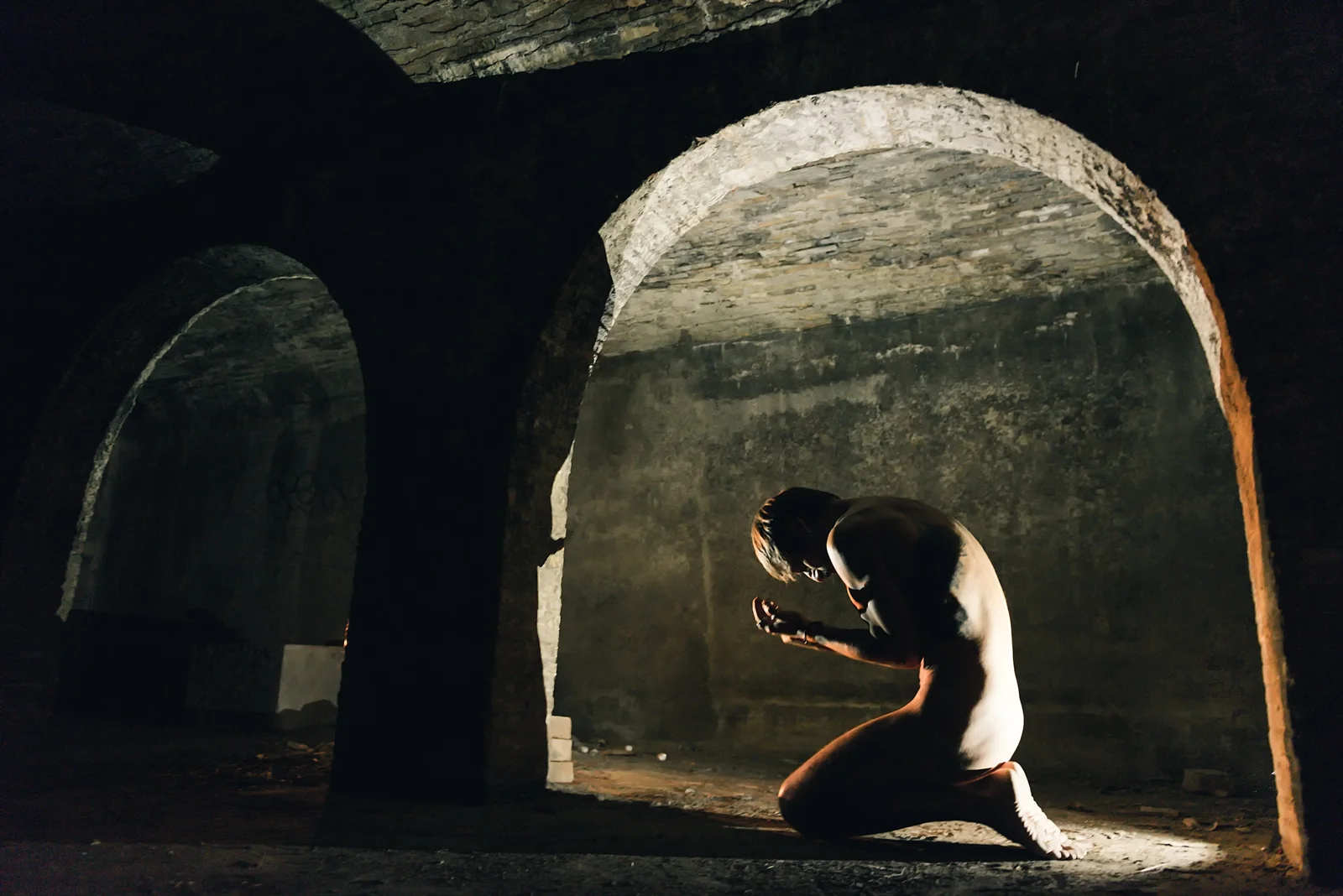 Fine-art nude figure kneeling and bowing inside an illuminated arch.