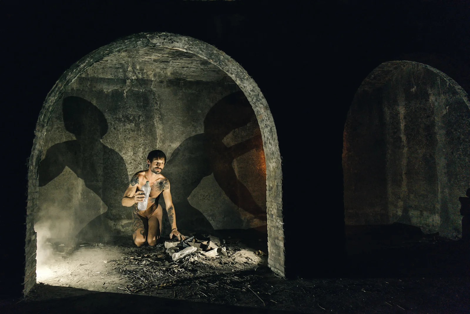 Wide view of a kneeling figure and shadows in a vaulted stone room.