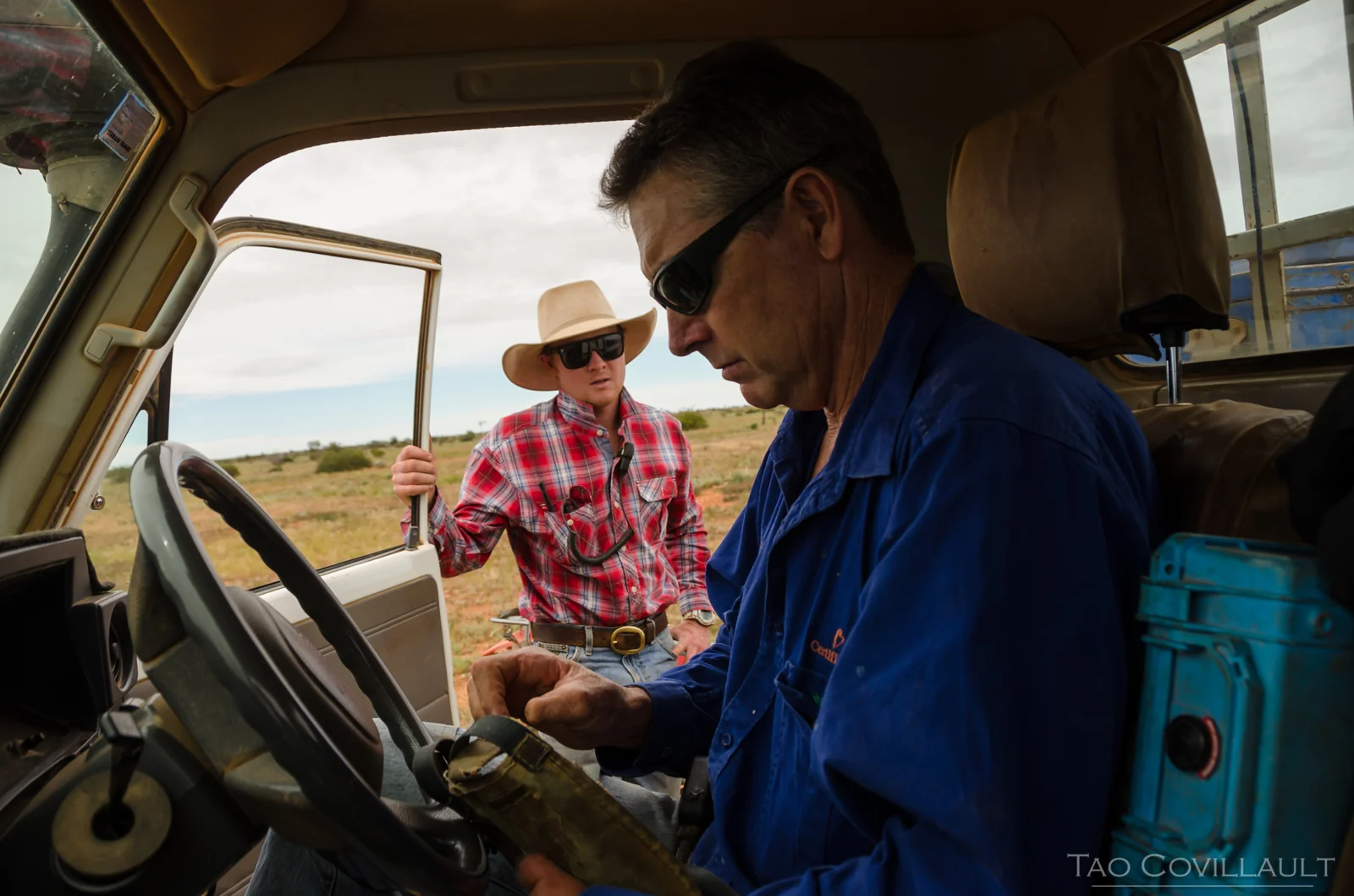 Man at the wheel of a farm vehicle with another stockman outside.