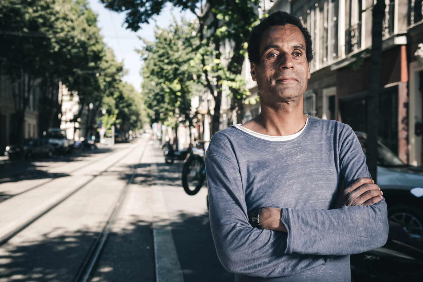 Man in a grey shirt standing with folded arms beside tram tracks.