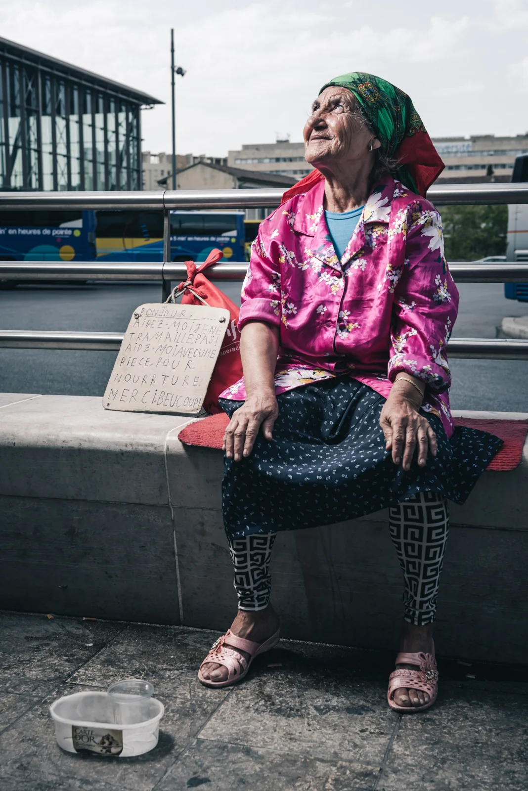 Woman in a pink jacket seated beside a bowl outside a station.